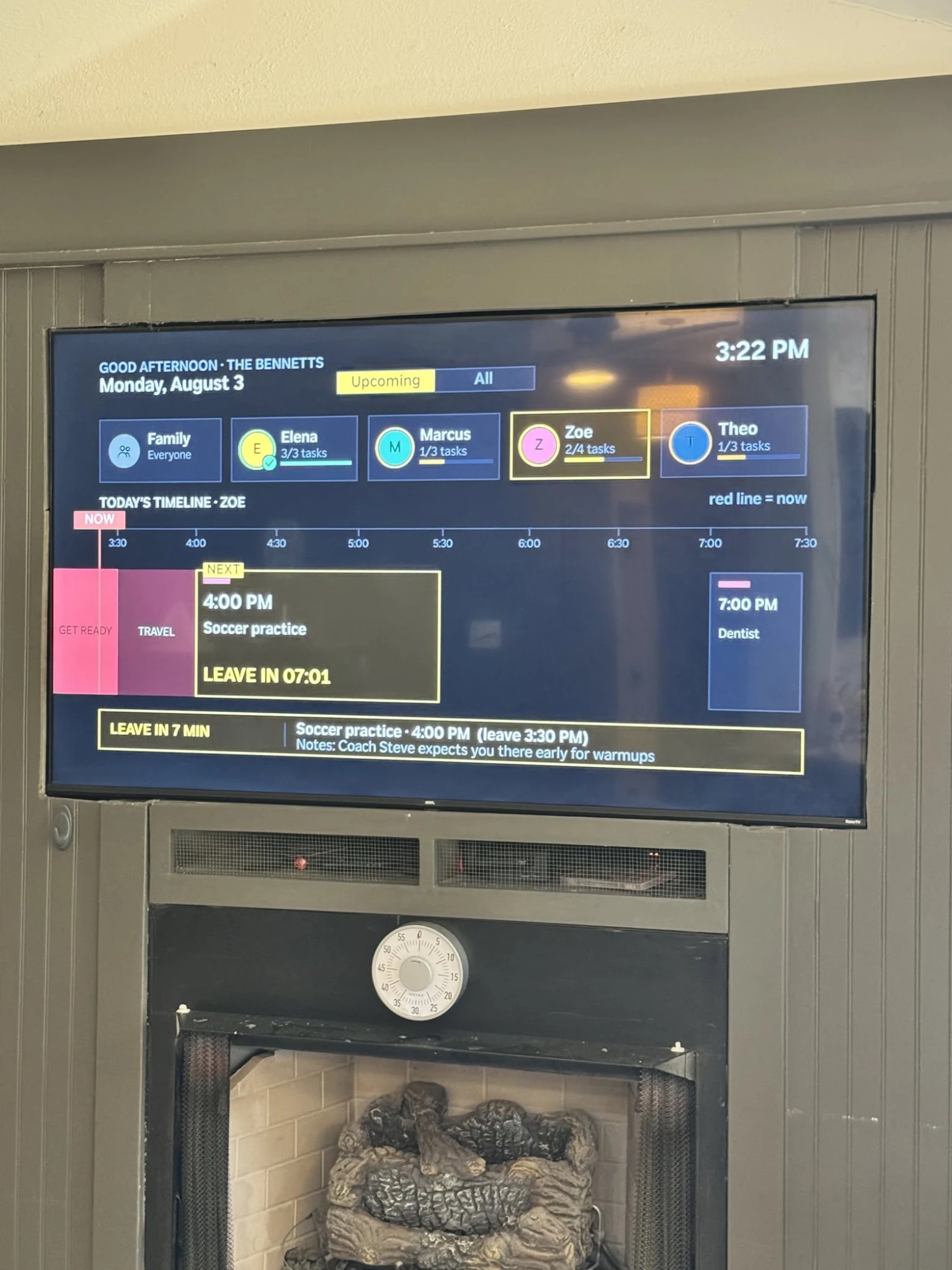A real wall-mounted TV above a fireplace running an early FamilyOrganization.app beta build. Zoe's timeline shows a GET READY block and a TRAVEL block before soccer practice at 4:00 PM, and the banner reads LEAVE IN 07:01.